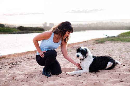 A woman kneels on the sand, engaging with her dog beside a calm river during sunset - Australian Stock Image