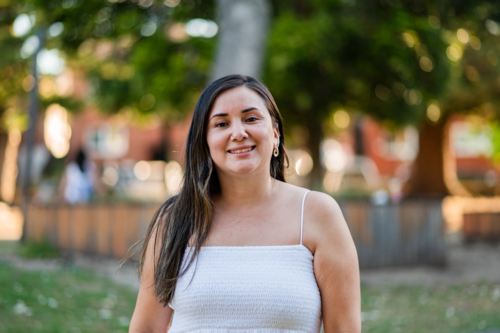 A woman stands in a park smiling while sunlight filters through the trees in the background - Australian Stock Image