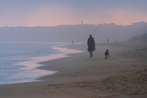 A woman walking with her dog on a foggy beach at sunset