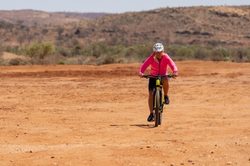A woman wearing a bright pink top riding a mountain bike through outback landscape - Australian Stock Image