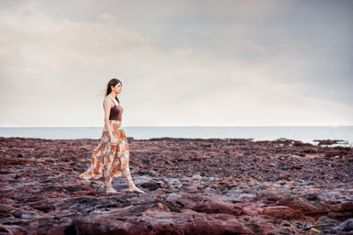 A woman wearing a long skirt walking on a rocky beach with ocean as the background - Australian Stock Image