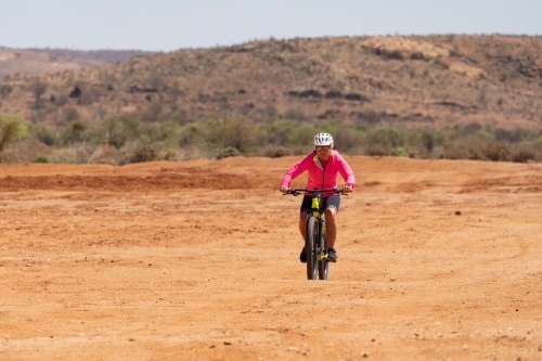 A woman wearing bright pink top riding a mountain bike through an outback landscape - Australian Stock Image