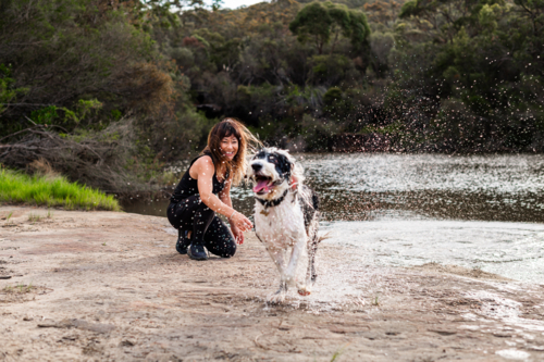 A woman with long hair enjoys quality time by the river, laughing as her dog splashes around. - Australian Stock Image