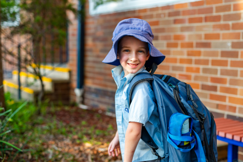 A young boy in a school uniform stands outside a primary school wearing a hat and smiling - Australian Stock Image