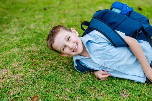 A young boy in school uniform is lying on the grass with a backpack while smiling - Australian Stock Image