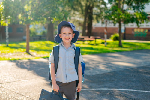 A young boy stands in the schoolyard, wearing a school uniform with a hat, wearing a backpack - Australian Stock Image