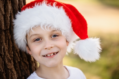 A young caucasian Australian boy wearing a Santa hat leaning against a tree and smiling at Christmas