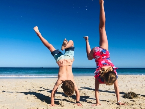 A young girl and boy doing handstands in swimming outfits on shoreline of beach on still summers day