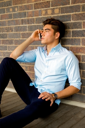 A young male sitting on the ground on a covered verandah talking on the phone - Australian Stock Image