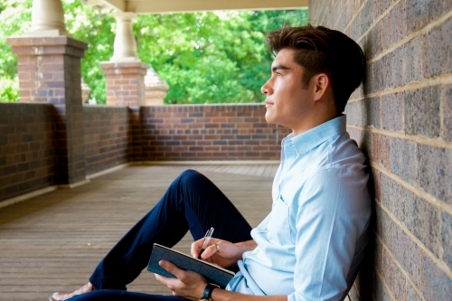 A young male sitting on the ground on a covered verandah with notebook - Australian Stock Image