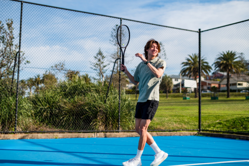 A young male tennis player smiles on a blue court amid greenery and palm trees - Australian Stock Image