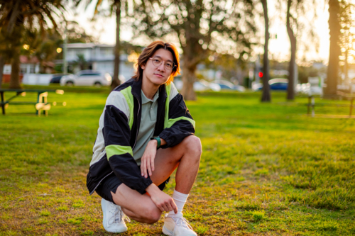 A young man crouches on the grass wearing casual sports clothes as the sun sets nearby - Australian Stock Image