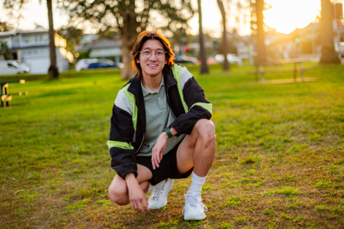 A young man kneels in a park at sunset wearing a relaxed outfit and smiling happily - Australian Stock Image
