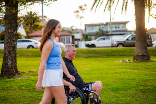 A young woman walks alongside a man in a wheelchair at a park, enjoying sunset together - Australian Stock Image