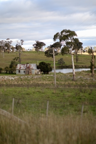 Abandoned work mans hut situated in a paddock by a dam full of water in the afternoon - Australian Stock Image