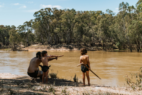 Aboriginal Australian father teaching children on banks of river - Australian Stock Image