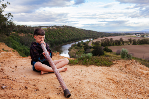 aboriginal boy playing didgeridoo in Australian landscape with view to Hunter River - Australian Stock Image