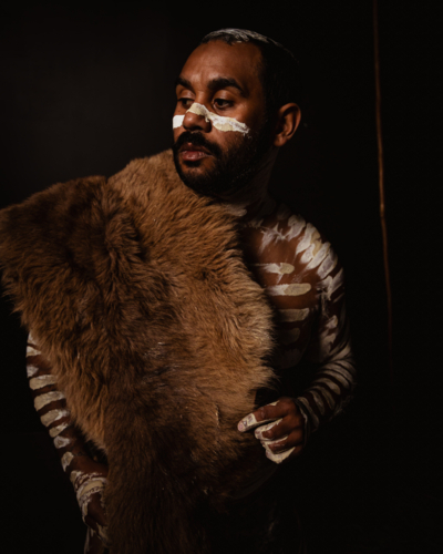 Aboriginal man wearing a fur cloak standing in dark background - Australian Stock Image