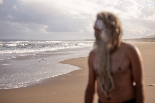 Aboriginal man with clay face paint standing on a beach focus on the water and sand