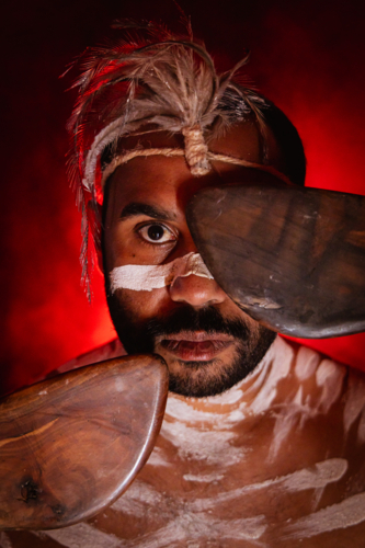 Aboriginal man with red background covering one eye with boomerang - Australian Stock Image