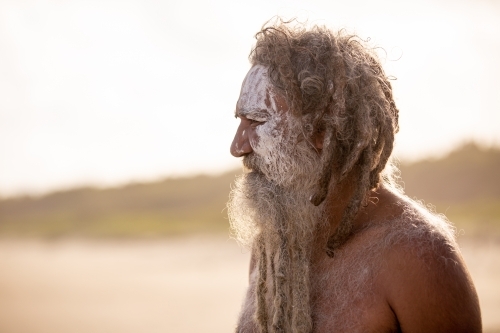Aboriginal middle aged man with clay face paint standing on a beach looking into distance