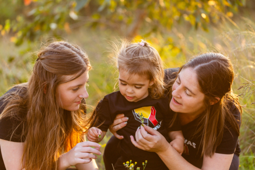 Aboriginal toddler playing outside in nature with big sister and mum looking at flowers - Australian Stock Image