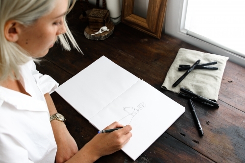 Above over the shoulder shot of young blonde woman drawing in sketch book with pens