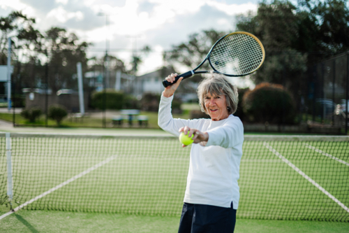 Active older woman preparing to hit a tennis ball outdoors while smiling - Australian Stock Image