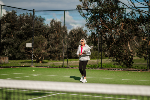 Active senior woman enjoying a game of tennis outdoors on a bright day, wearing sunglasses. - Australian Stock Image