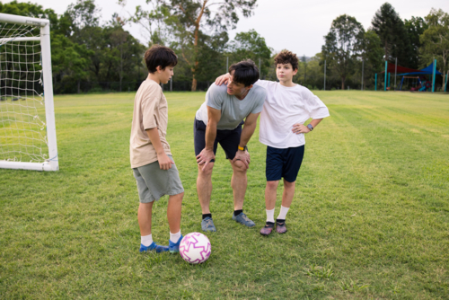 Adult man with two young boys in a huddle on grassy soccer field near goalpost - Australian Stock Image