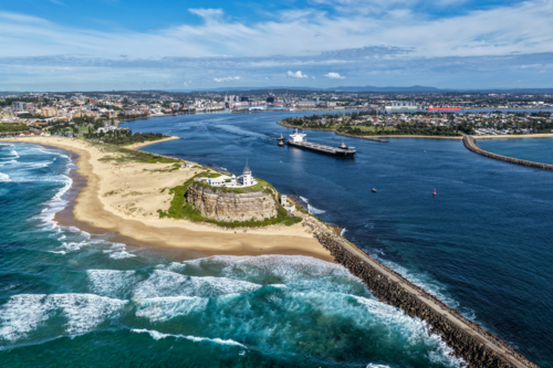 Aerial drone view of Nobbys Lighthouse and Nobbys Beach with a large cargo ship being escorted. - Australian Stock Image