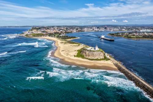 Aerial drone view of Nobbys Lighthouse and Nobbys Beach with a large cargo ship being escorted. - Australian Stock Image