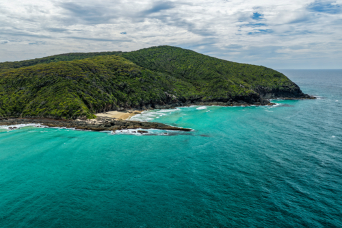 Aerial drone view of Sharkies Beach featuring a rugged rocky coastline and a tree covered mountain. - Australian Stock Image