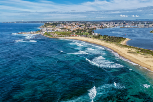 Aerial drone view of waves rolling onto the sandy shoreline at Nobbys Beach with Newcastle city - Australian Stock Image