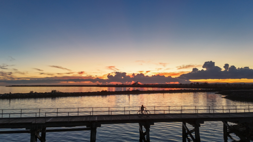 Aerial of silhouette of person on bike riding along jetty at sunset - Australian Stock Image
