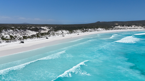 Aerial over the ocean of cars parked along remote beach with crystal blue water and white sand - Australian Stock Image