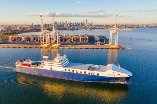 Aerial view of a cargo ship cruising past large gantry cranes on a dock - Australian Stock Image