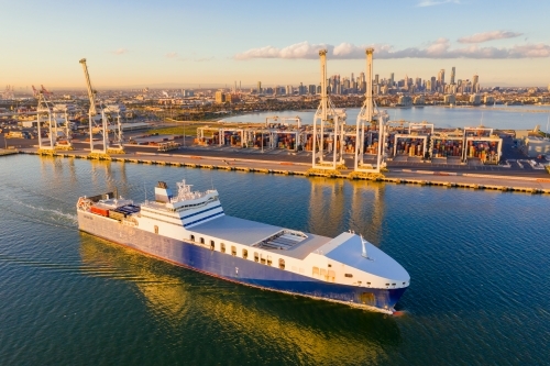 Aerial view of a cargo ship cruising past large gantry cranes on a dock - Australian Stock Image