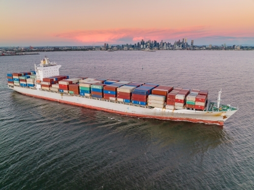 Aerial view of a cargo ship leaving port at sunset, fully laden with containers - Australian Stock Image