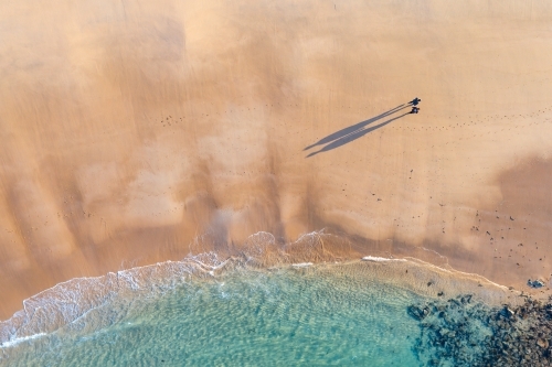 Aerial view of a couple walking on a wide sandy beach castling long shadows - Australian Stock Image