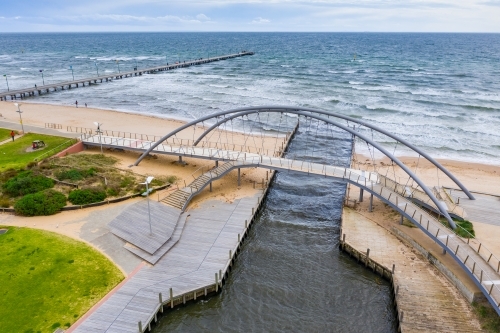 Aerial view of a modern arched walking bridge over a marina channel flowing out to sea.