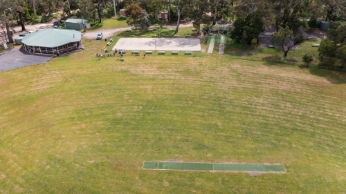 Aerial view of a people gathered at a grassy oval with a cricket pitch and clubrooms