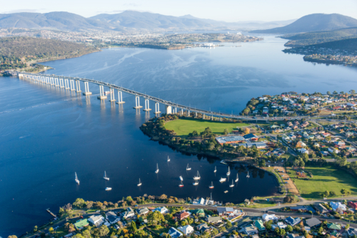 Aerial view of bridge crossing wide river with sailboats and suburban houses below - Australian Stock Image