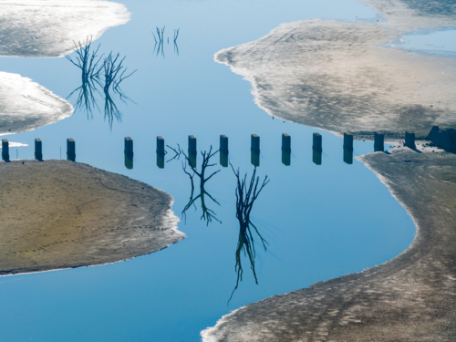 Aerial view of dead trees and derelict bridge pillars crossing a still blue stream - Australian Stock Image