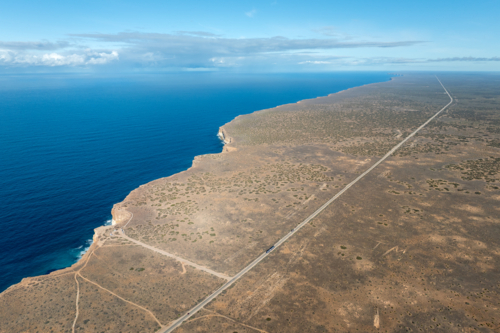 Aerial view of dramatic limestone cliffs dropping to deep blue ocean beside a straight outback road - Australian Stock Image