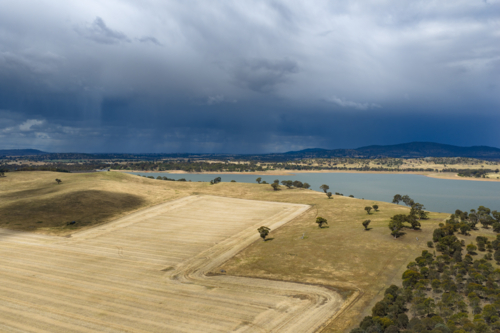 Aerial view of dry harvested farmland alongside a reservoir with rain falling from dark clouds above - Australian Stock Image