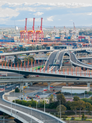Aerial view of intersecting and curving freeways with shipping cranes rising in the distance - Australian Stock Image