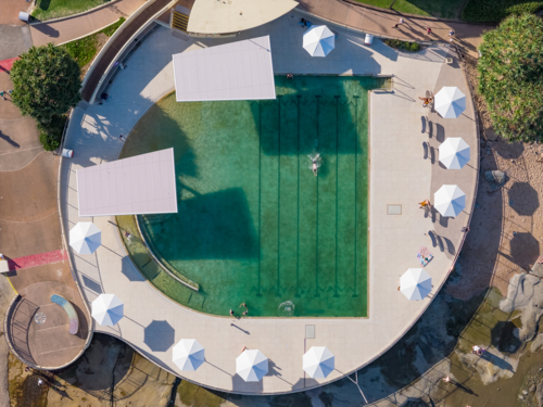Aerial view of people swimming laps in a coastal pool with umbrellas alongside - Australian Stock Image