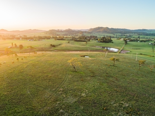 Aerial view of scattered gum trees in green farm paddock with dam and distant hills at sunset - Australian Stock Image