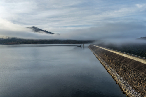 Aerial view of the Glenbawn Dam wall and surrounding landscape with fog rolling through the valley - Australian Stock Image